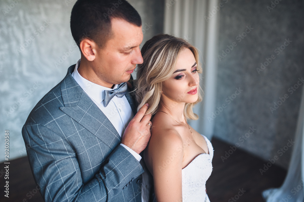 Beautiful and modern newlyweds stand in a gray studio. A stylish groom adjusts the hair to a sweet bride with curly hair. Portrait of a newlywed couple. Beautiful and modern newlyweds stand in a gray studio. A stylish groom adjusts the hair to a sweet bride with curly hair. Portrait of a newlywed couple.