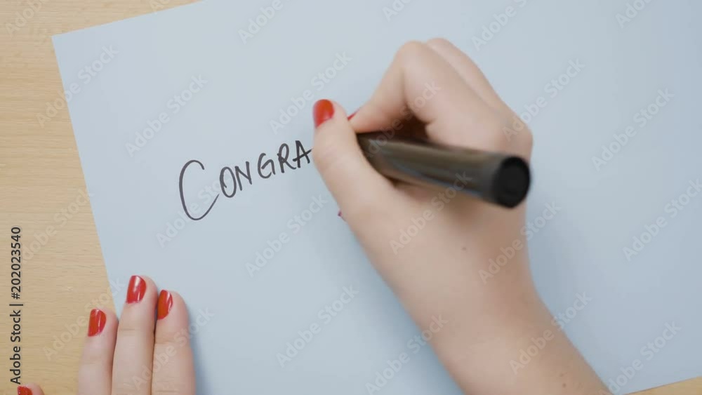 Female hands writing congratulations on a blue paper using a black marker Female hands writing congratulations on a blue paper using a black marker