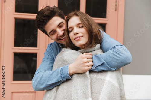 Photo of happy couple in love standing outside cafe, while man hugging and wrapping his woman in warm blanket