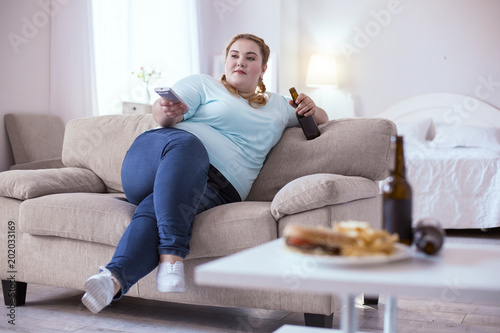 Bad nutrition. Stout red-head woman drinking beer while watching television photo