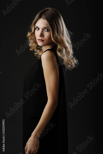 Beautiful blonde hair woman in black dress looking back while posing in the studio on black background. Fashion portrait