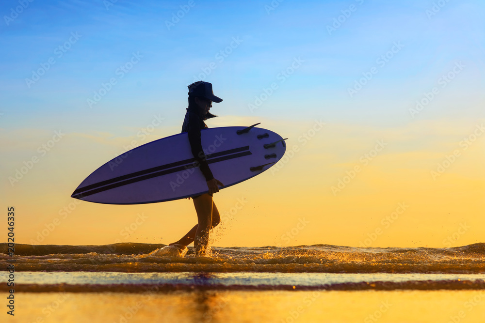 Silhouette of woman holding surfboard beside walking on the edge of sea wave, splash of water kicking while walking through, returning home stattion after surfed in the sea at sunset light