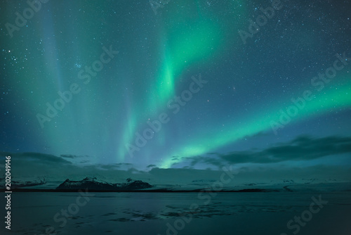 Northern lights aka Aurora Borealis glowing on sky in Jokulsarlon glacier lagoon with snow capped mountains in background at night in Iceland