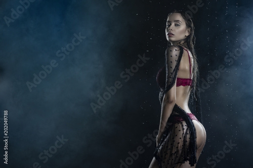 Young wet slim girl wearing a red lingerie and black translucent veil posing sideways in rain water drops in a studio shows her beautiful butt on black in a theatrical smoke