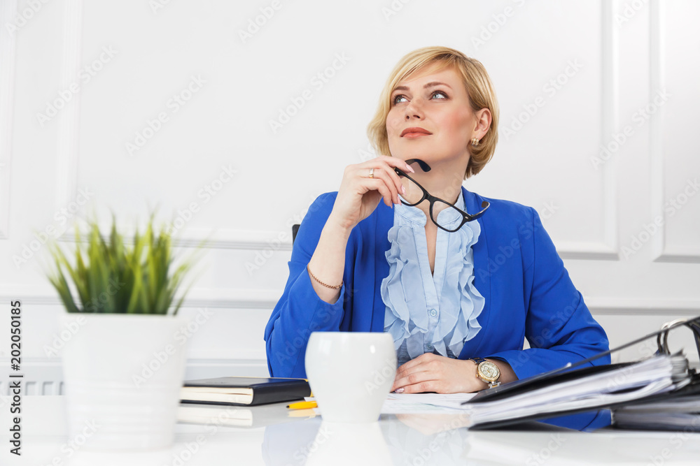 Attractive blonde businesswoman wears blue jacket holds eyeglasses while thinking, boss at workplace manage with documents, white office room Attractive blonde businesswoman wears blue jacket holds eyeglasses while thinking, boss at workplace manage with documents, white office room