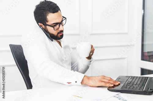 Handsome bearded businessman wears white shirt work on laptop and talk by phone in white office interior