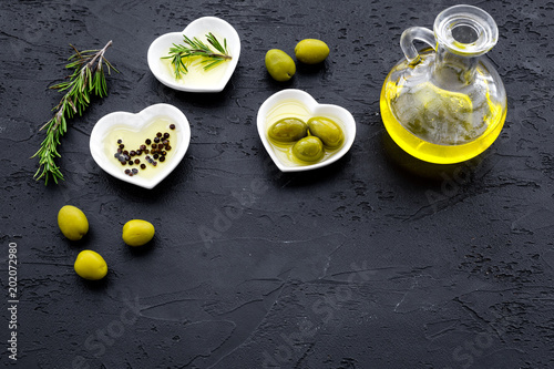 Fresh olive oil in glass jar near green olives and branch of rosemary on black background top view space for text