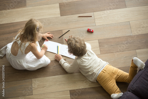 Kids sister and brother playing drawing together on wooden warm floor in living room, creative children boy and girl having fun at home, siblings friendship, underfloor heating concept, top view