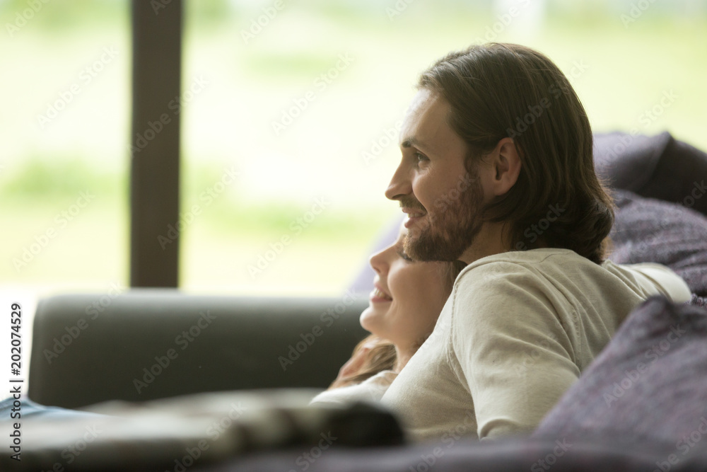 Young beautiful loving couple relaxing on sofa together, happy millennial family sitting on couch on cozy warm leisure day, husband and wife resting bonding at home enjoying weekend, side view Young beautiful loving couple relaxing on sofa together, happy millennial family sitting on couch on cozy warm leisure day, husband and wife resting bonding at home enjoying weekend, side view