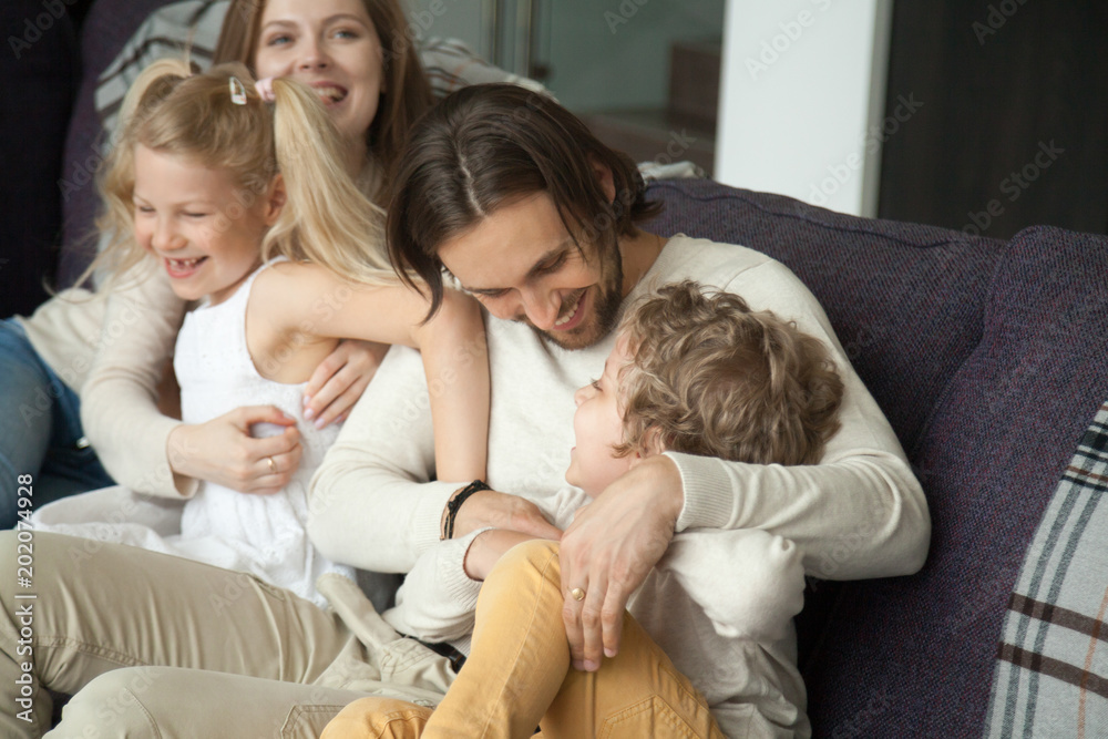 Happy parents and children laughing having fun tickling on sofa, cheerful mom and dad embracing kids playing with son and daughter on couch, funny weekend activity and good family relations concept