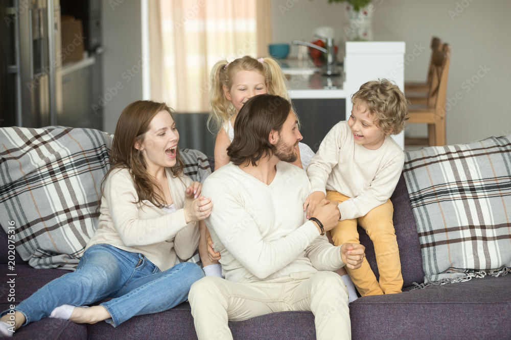 Kids and parents laughing having fun together sitting on sofa, smiling excited children son daughter playing with mom and dad at home, playtime funny weekend activity, happy family lifestyle concept