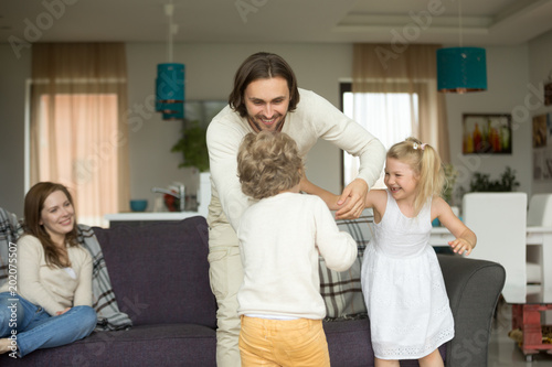 Happy young dad having fun with kids at home, smiling daddy playing with children son and daughter in living room while mom relaxing on sofa, father laughing enjoying weekend activities with family