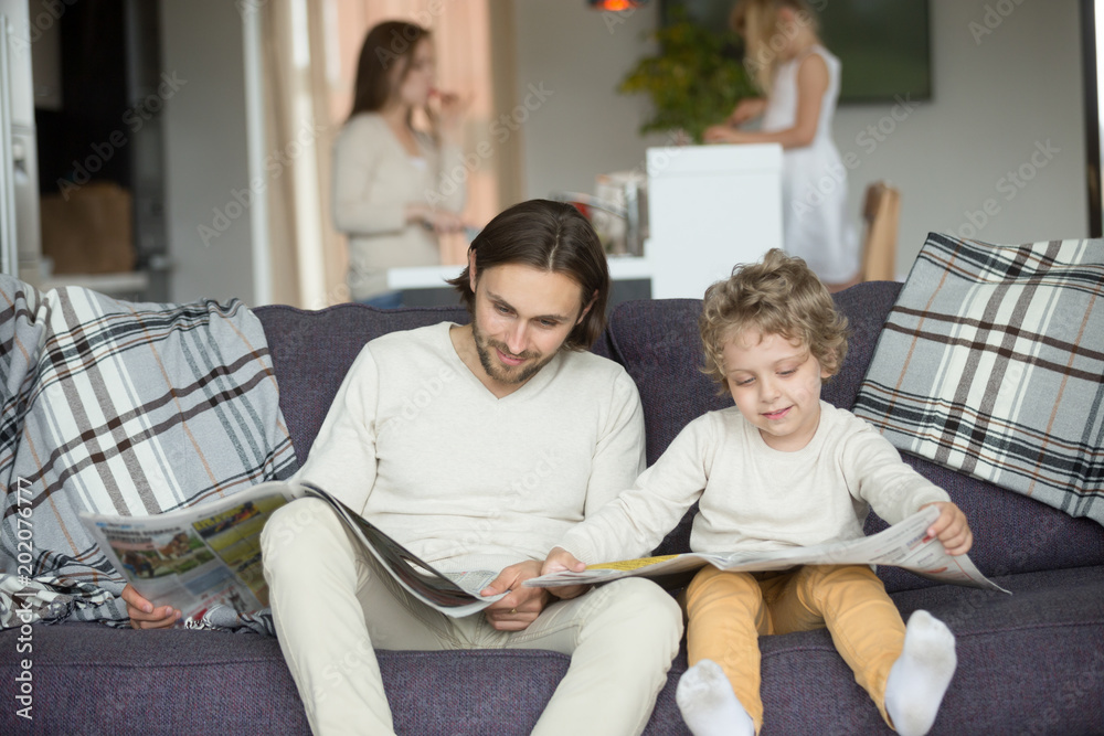 Obraz premium Little son pretending reading newspaper sitting on couch with father, cute funny kid imitating dad to be alike spending weekend with family, child boy learning to read discussing news with daddy
