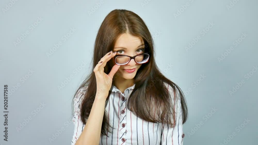 Attractive Young Girl in White Shirt with Red and Blue Stripes Looking at the Camera with Interest Over Her Eyeglasses with Charming Smile in Studio on Grey Background. Attractive Young Girl in White Shirt with Red and Blue Stripes Looking at the Camera with Interest Over Her Eyeglasses with Charming Smile in Studio on Grey Background.