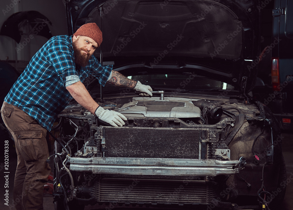 A bearded expert mechanic dressed in a uniform, standing with crossed arms against a car in the garage.