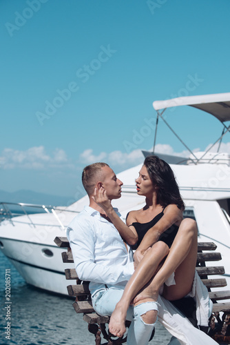 Young passionate couple sit on bench at dock near boat and hugs, sunny summer day. Woman and man in fashionable clothes full of desire near luxury yacht. Honeymoon concept. Couple in love on honeymoon