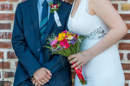 Bride and groom together in front of brick wall. Bride holding wedding bouquet of colorful flowers. Groom with crossed, folded arms and red rose boutonniere on jacket.