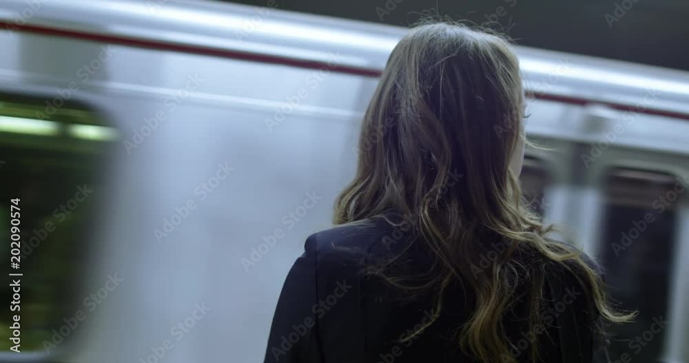 OTS closeup, smartly dressed business woman watches as subway train pulls into station in front of her. Hand-held, slow motion 4K 60fps. Logos and other people obscured by motion blur