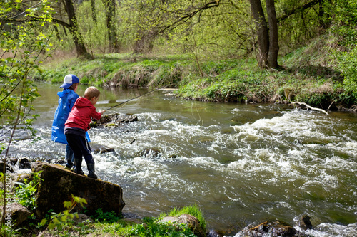 two boys brothers, dressed in red and blue raincoat, are fishing together on the mount river