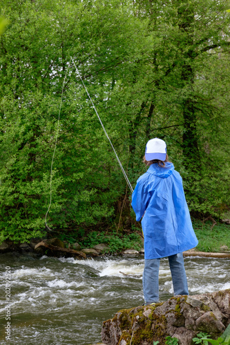 boy, dressed in blue waterproof raincoat and cap, fishing on the mountain streamy river © Kiryl Lis