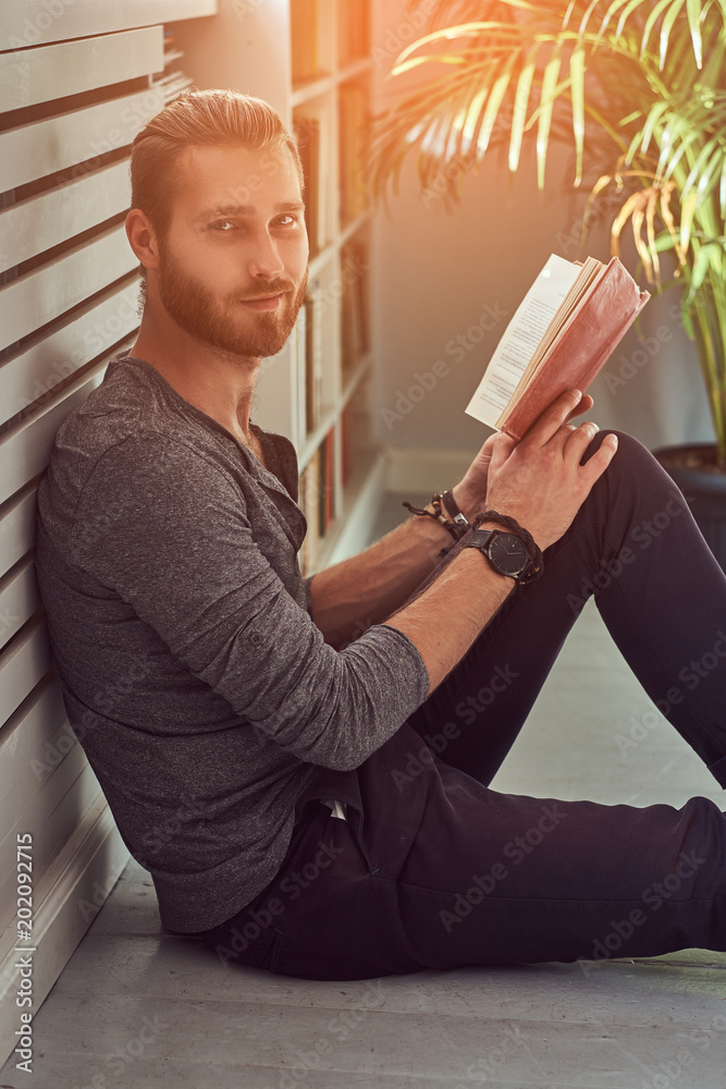 Fototapeta premium Handsome stylish redhead student in casual clothes, sitting on a floor at a home and holds a book, looking at a camera.