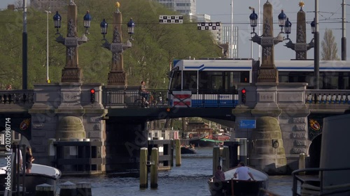 Canal boats cruising and a tram is passing over the Amstelbrug from the Amstel river on a sunny day in Amsterdam. 4K photo