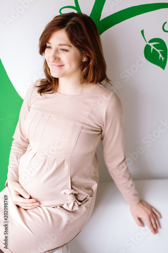 pregnancy, happiness, childbirth concept. against the wall with greeny patterns of leaves there is a smiling woman with midle haircut, light shining complexion, dressed in gorgeous cremy dress photo