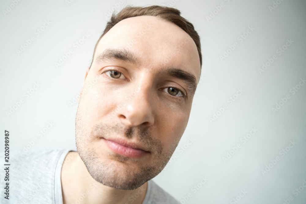 Obraz premium Portrait of a young handsome fashionable guy with a stubble and a smile on his face in a gray shirt on a light background. Wide angle, very large portrait