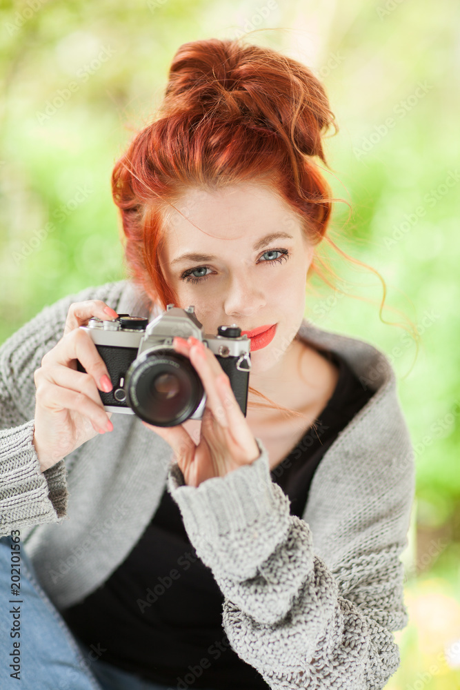 Beautiful young woman with red hair sitting in the garden taking pictures with old, analog camera Beautiful young woman with red hair sitting in the garden taking pictures with old, analog camera
