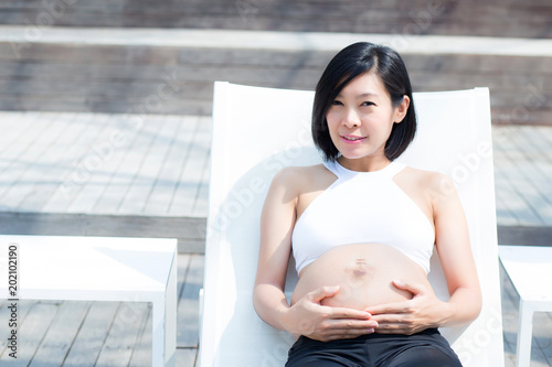 Portrait of beautiful pregnant asian young woman relax in the park, girl with belly sitting at swimming pool for wellness, tummy girl, health care concept. photo