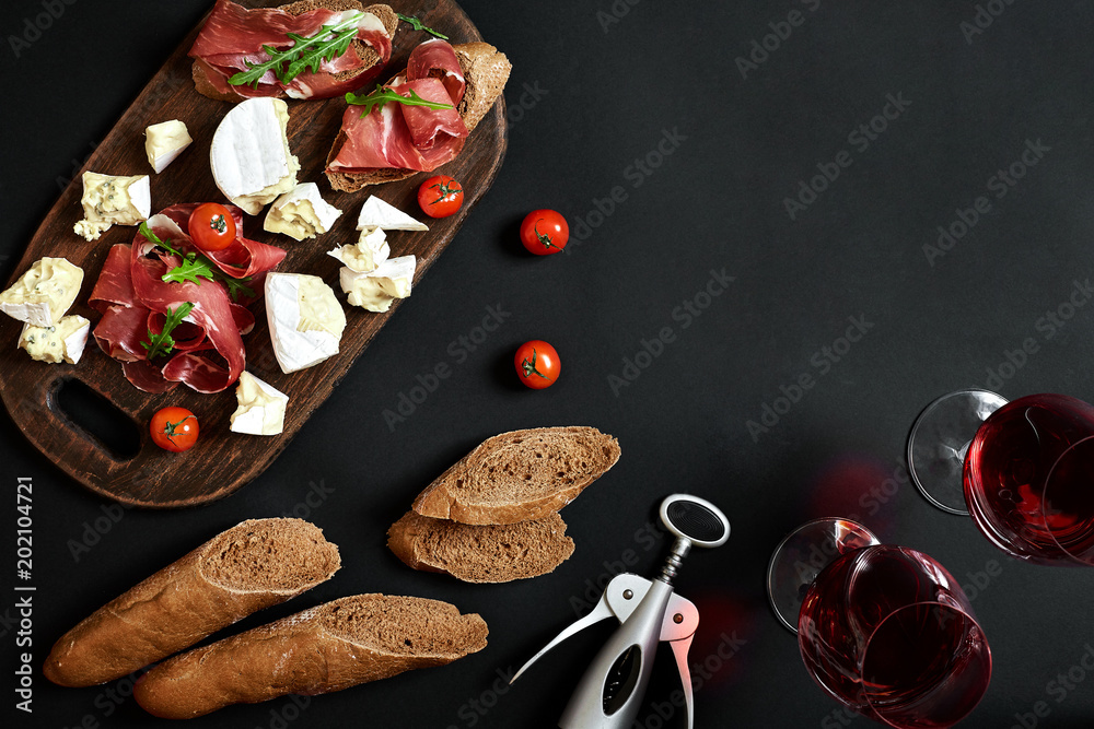 Prosciutto, salami, baguette slices, tomatoes and nutson rustic wooden board, two glasses of red wine over black background