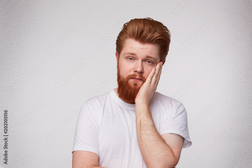 Indoor shot of bearded ginger male feels tired, keeps his hand on cheek, looks forworn and aweary. Isolated over white studio wall Indoor shot of bearded ginger male feels tired, keeps his hand on cheek, looks forworn and aweary. Isolated over white studio wall