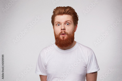 Indoor shot of shoked stupefied young ginger male looks with frightened expression, isolated over white background