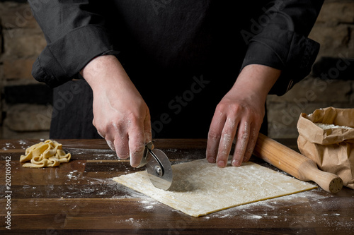 Hands of a man close-up, cut into slices dough for pasta, on a wooden table on the background of a brick wall.