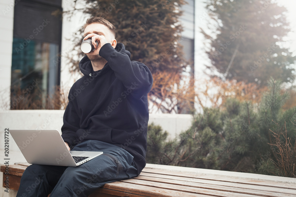 Young man holding laptop on kneesand drinking coffee from black cup. Young man holding laptop on kneesand drinking coffee from black cup.