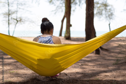 Young teenager girl with long hair but tie it up is sitting on yellow hammock facing with the sea on her relaxing holiday travel