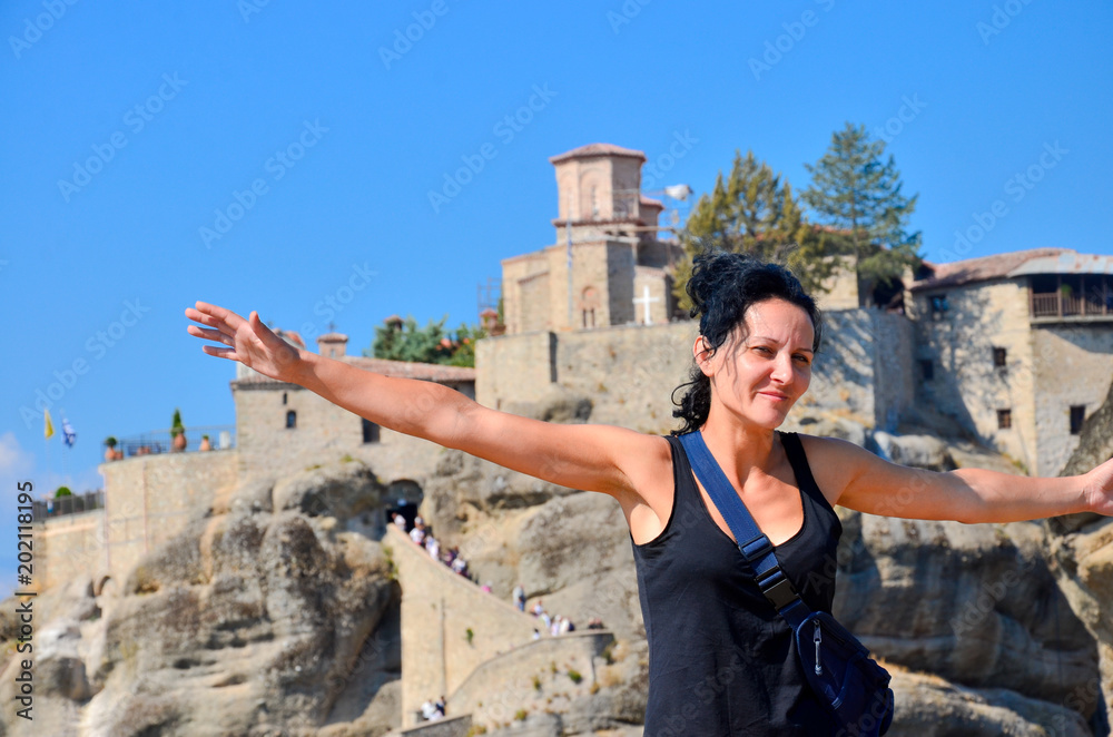 Young happy woman stands back to the rock with raised hands and monastery at background. Traveler enjoying the landscape with mountain, Monasteries of Meteora, Greece