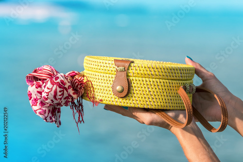 Woman hands with fashionable stylish rattan bag and silk scarf on the ocean background. Tropical island of Bali, Indonesia. Rattan handbag and silk scarf. photo