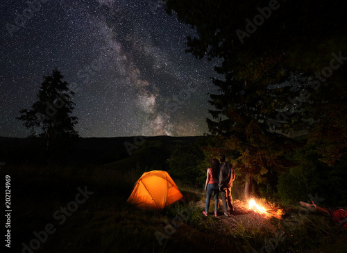 Rear view of young couple tourists enjoying the starry sky with a bright Milky way under the mighty trees near the campfire and orange illuminated tent in mountains. Romantic night camping near forest