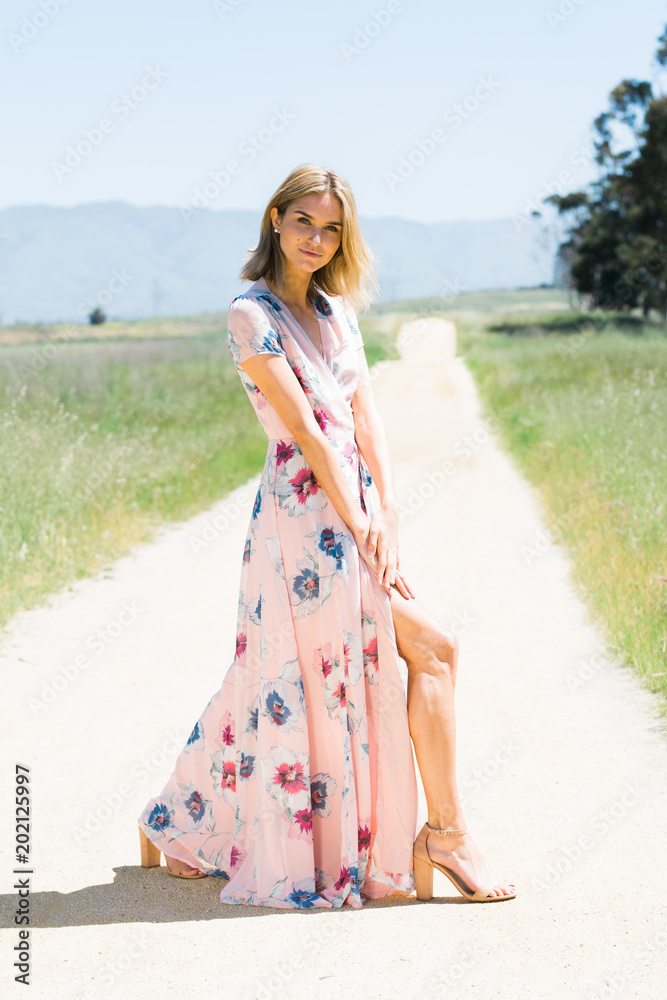 Attractive young blonde Caucasian woman in flowing pink floral print dress posing outside runway style on empty dirt road Attractive young blonde Caucasian woman in flowing pink floral print dress posing outside runway style on empty dirt road