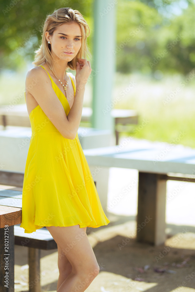 Attractive young blonde Caucasian woman in thin yellow sun dress posing outside on wooden table