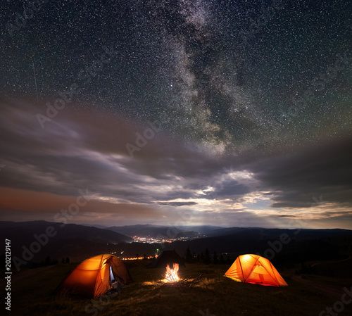 Camping illuminated orange tents on the top of mountain at night under starry sky and milky way on the background of the luminous town which is in the valley. Between the tents lit a bonfire