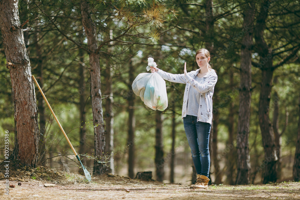 Fototapeta premium Young woman cleaning rubbish holding trash bags and showing stop gesture with palm in park on green background. Problem of environmental pollution. Stop nature garbage, environment protection concept.