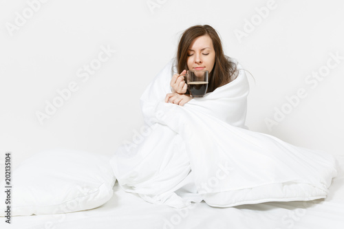 Young tired brunette woman sitting in bed with cup of coffee, white sheet, pillow, wrapping in blanket on white background. Female spending time in room. Rest, relax, good mood concept. Copy space.