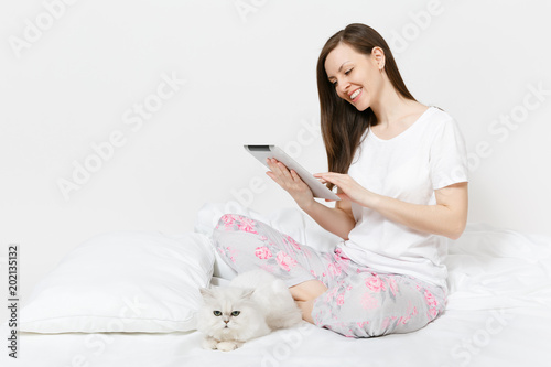 Young woman sitting in bed with white cute Persian silver chinchilla cat isolated on white background. Beauty female with tablet pc computer spending time in room. Rest, relax, good mood concept.