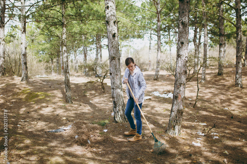 Young woman in casual clothes cleaning rubbish using rake for garbage collection in littered park or forest. Problem of environmental pollution. Stop nature garbage, environment protection concept.