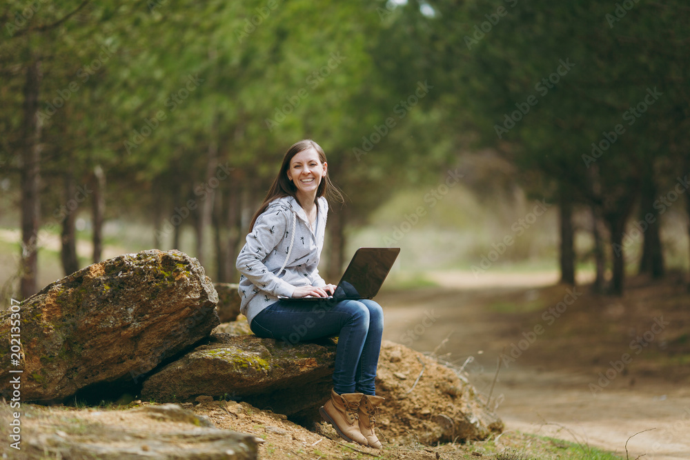 Young laughing successful smart business woman or student in casual clothes sitting on stone using laptop in city park or forest working outdoors on green blurred background. Mobile Office concept. Young laughing successful smart business woman or student in casual clothes sitting on stone using laptop in city park or forest working outdoors on green blurred background. Mobile Office concept.