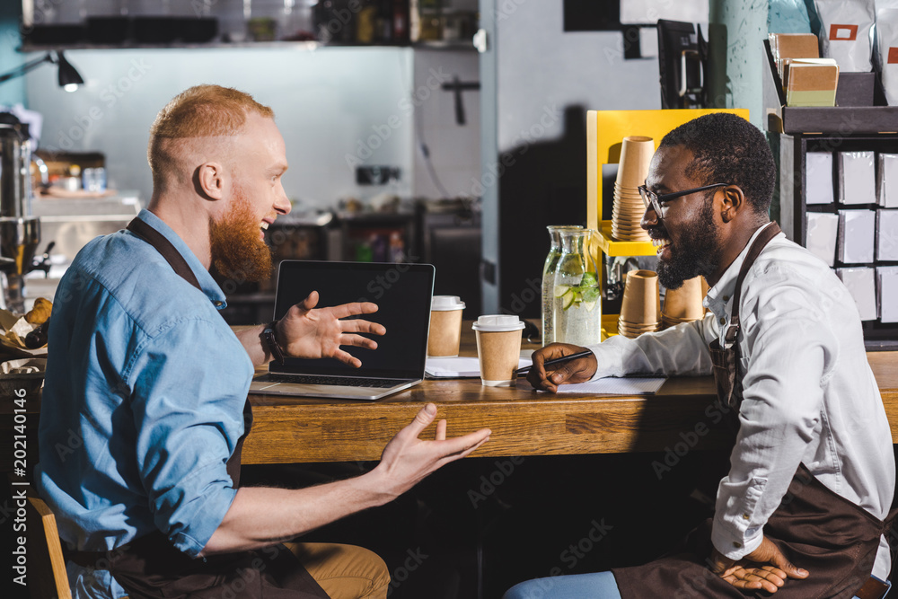 smiling young multicultural owners of coffee shop talking and sitting at table with laptop and coffee cups smiling young multicultural owners of coffee shop talking and sitting at table with laptop and coffee cups