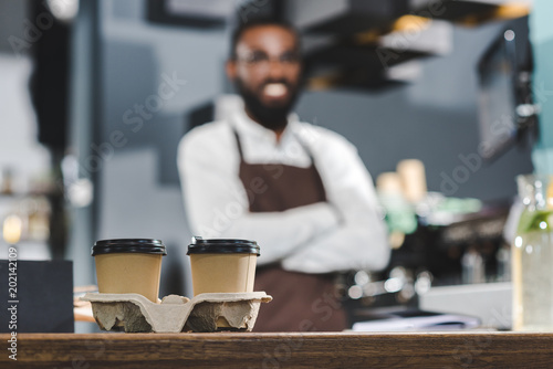 close-up view of two paper cups with coffee and smiling african american barista standing with crossed arms behind