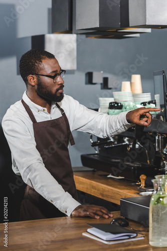 handsome young african american barista in eyeglasses pointing at screen while working in cafe